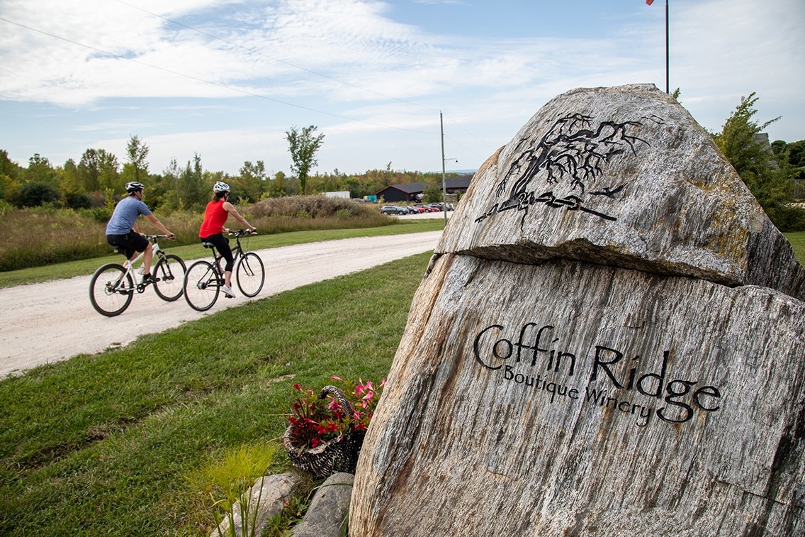 A large wooden stone with engraved lettering reads Coffin Ridge Boutique Winery. In the background two cyclists ride a gravel path.