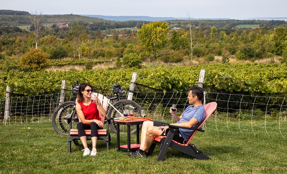 A couple sit beside vines drinking from small glasses.
