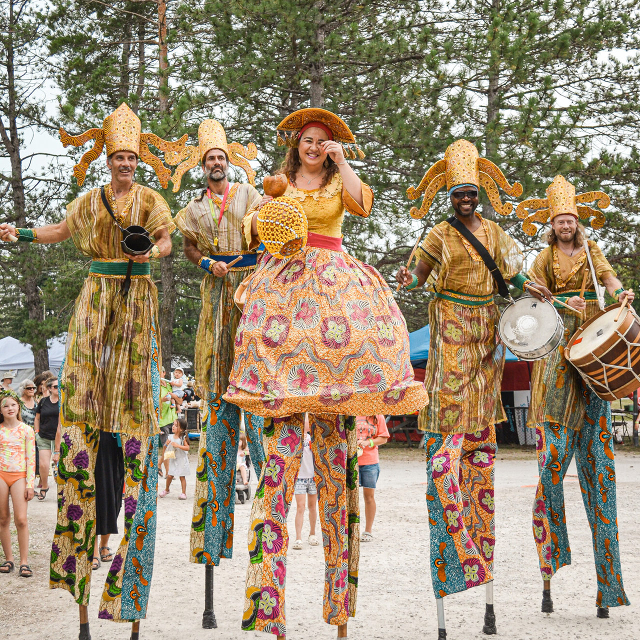 Five stilt performers in ornate, colorful costumes lead a festive outdoor parade among trees and onlookers.