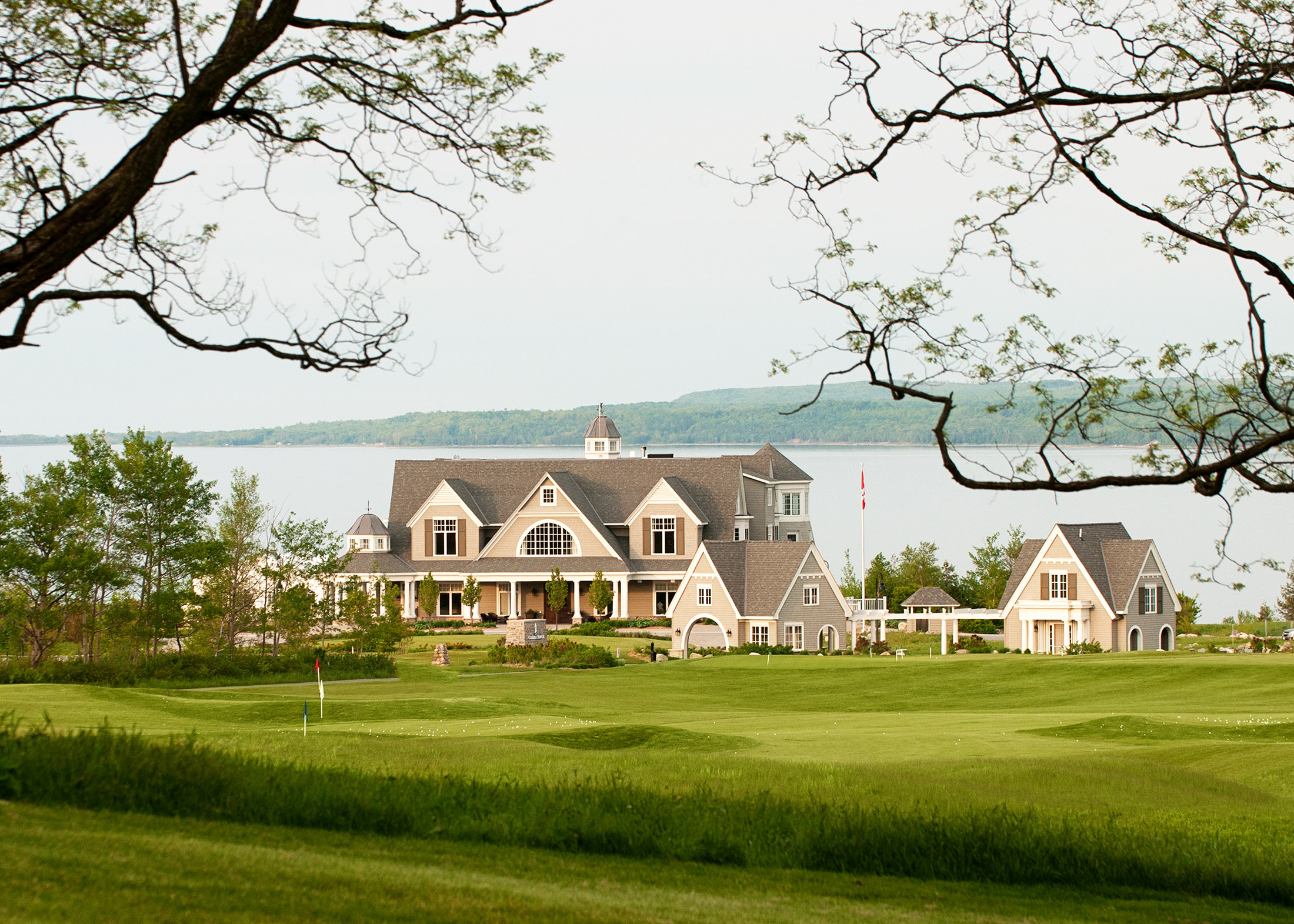 Lakeside clubhouse-style buildings surrounded by green golf course and framed by tree branches.