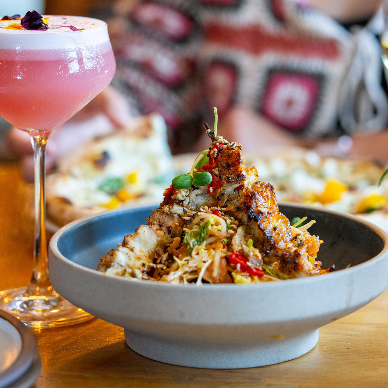 Crispy, spice-topped bites in a ceramic bowl beside a pink cocktail on a restaurant table.