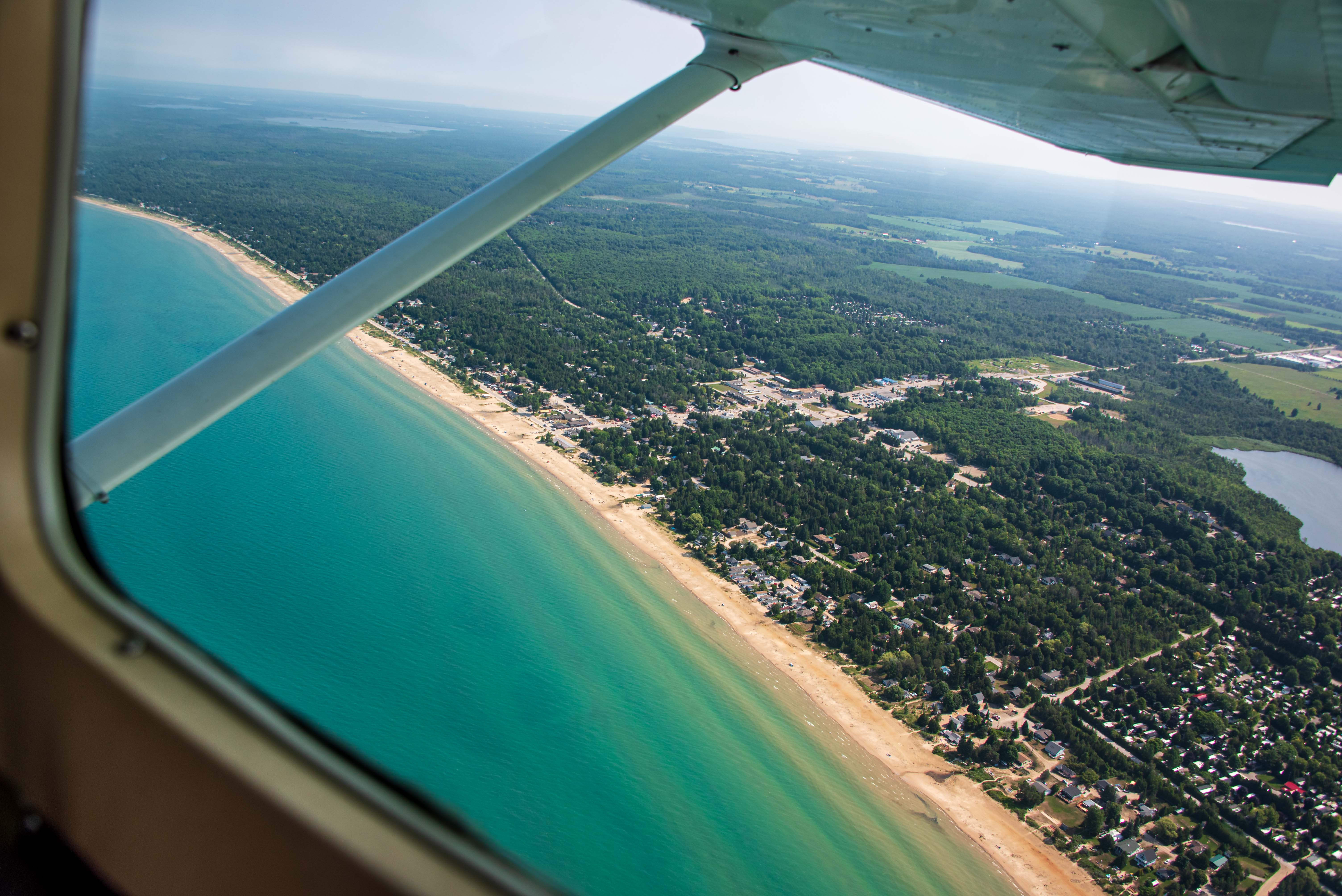 Looking out the window of a small aircraft toward a long sandy beach. Turquoise water, a sandy beach, a small downtown, cottages and trees are seen below.