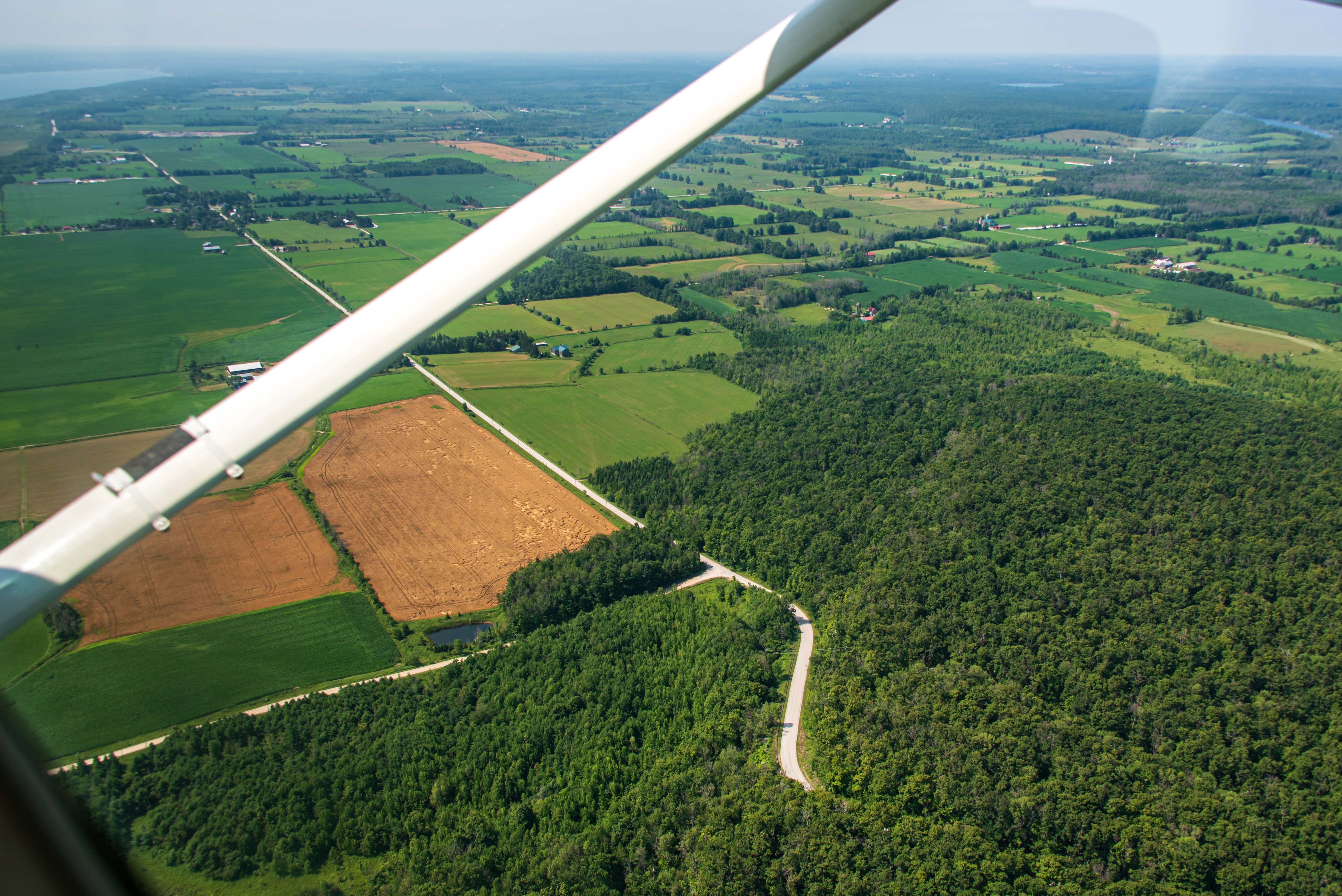 View from a small aircraft above a rural area. Forest, farmland, and a winding road are seen from above.