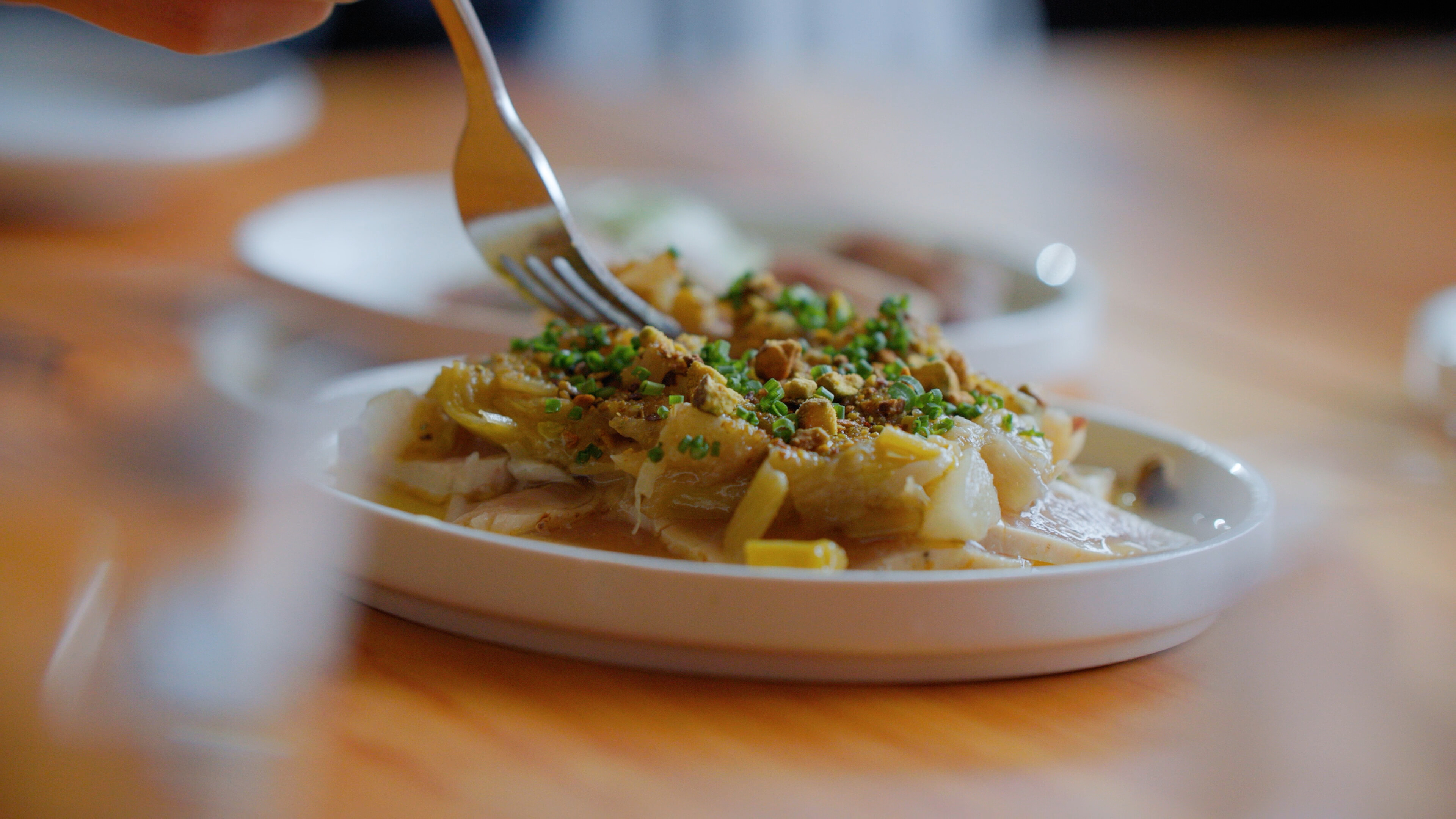 Fork cutting into a plated dish topped with herbs and crunchy garnish on a wooden table.