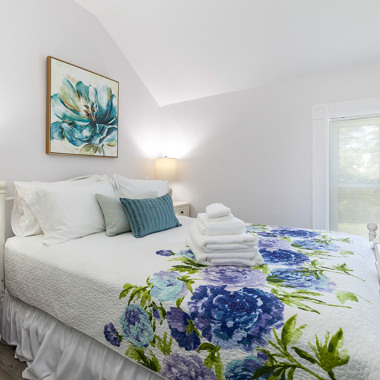 Bright, cozy guest room with a neatly made bed, floral quilt, folded towels, and soft natural light.