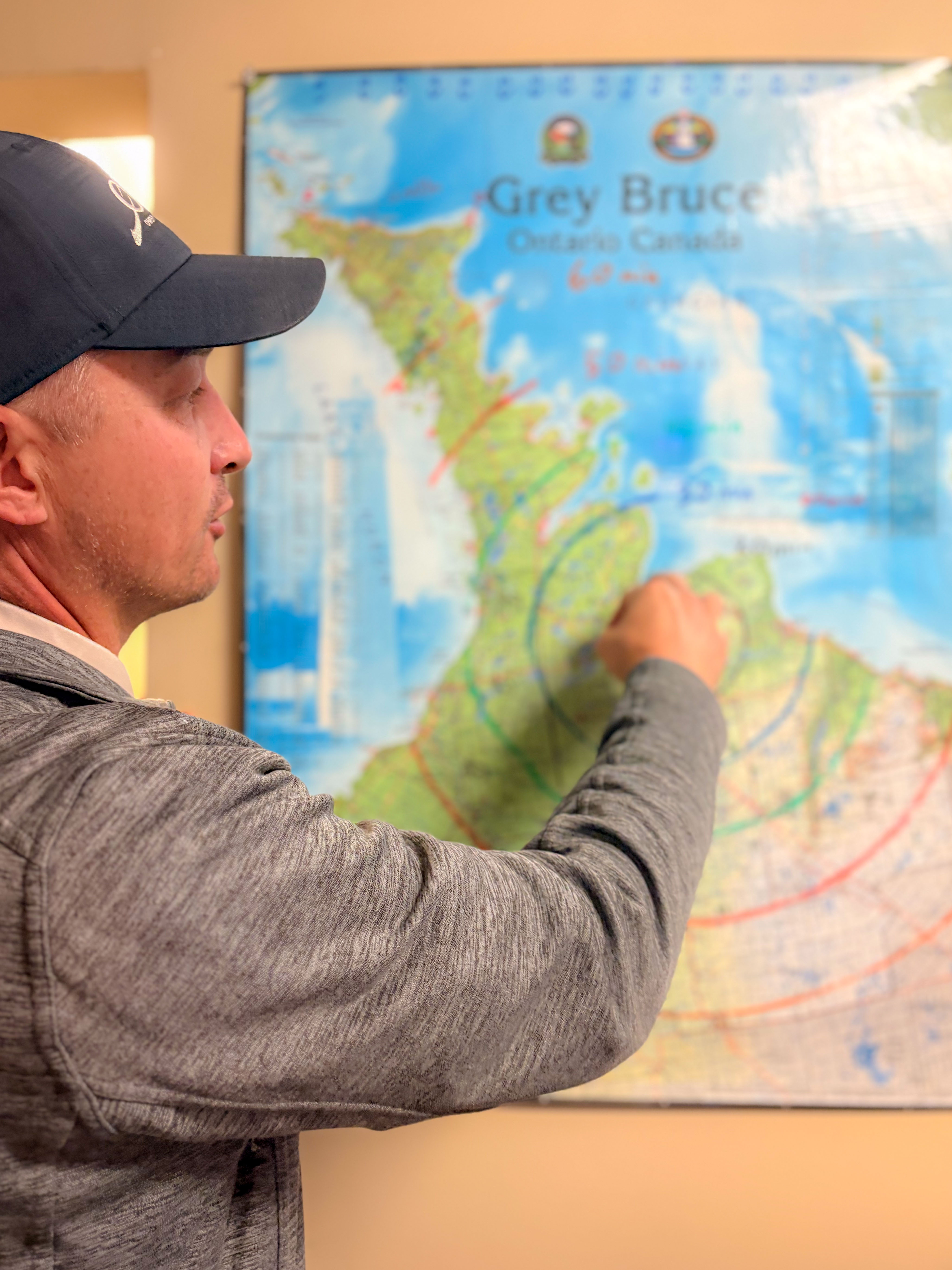 A man wearing a ballcap points at a map of Grey and Bruce counties showing flight paths.