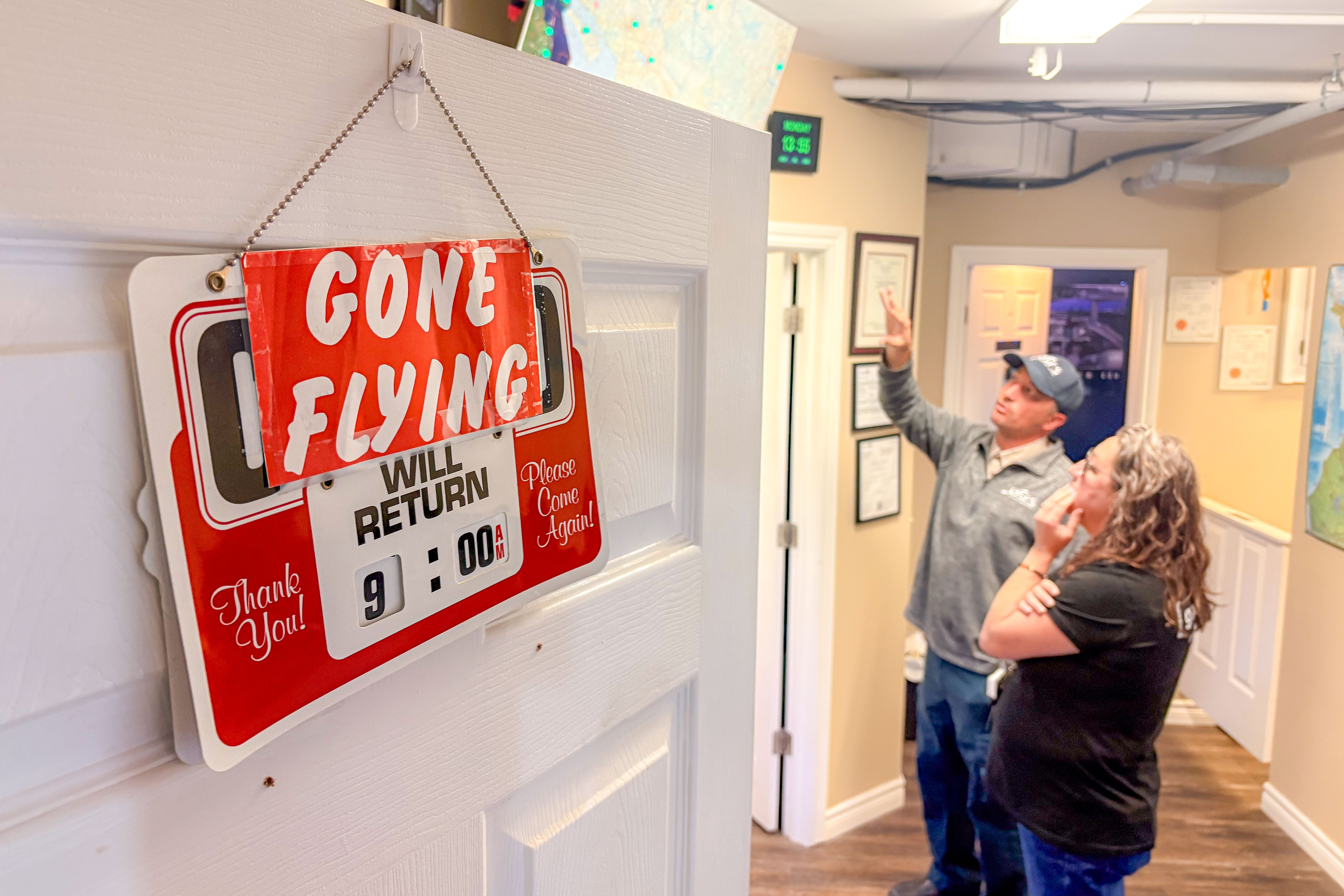 A "Gone Flying" sign hangs from an open door. Beyond, two people stand speaking in a well lit room.