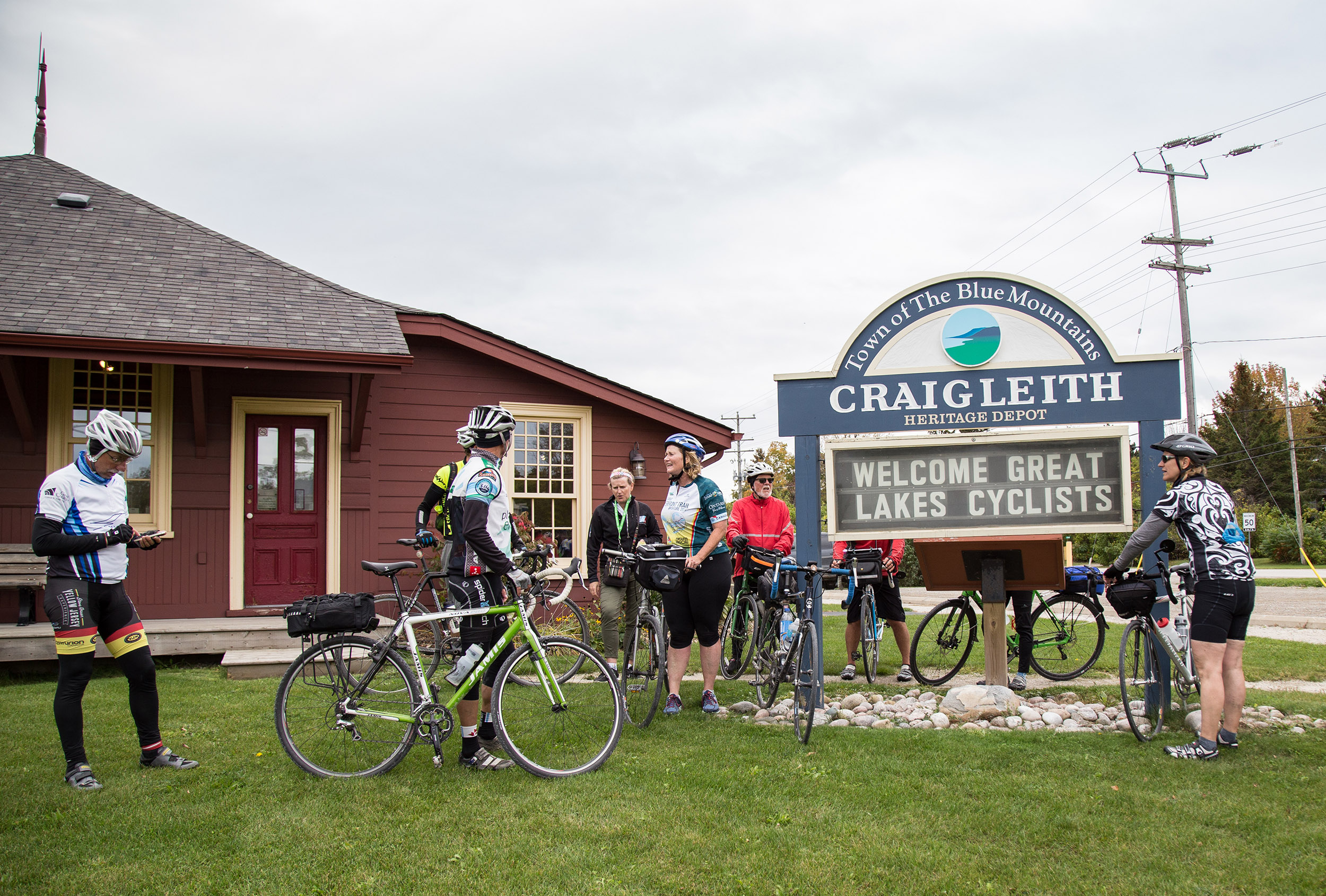 Cyclists with their bikes gather beside a heritage building and a sign welcoming Great Lakes cyclists.