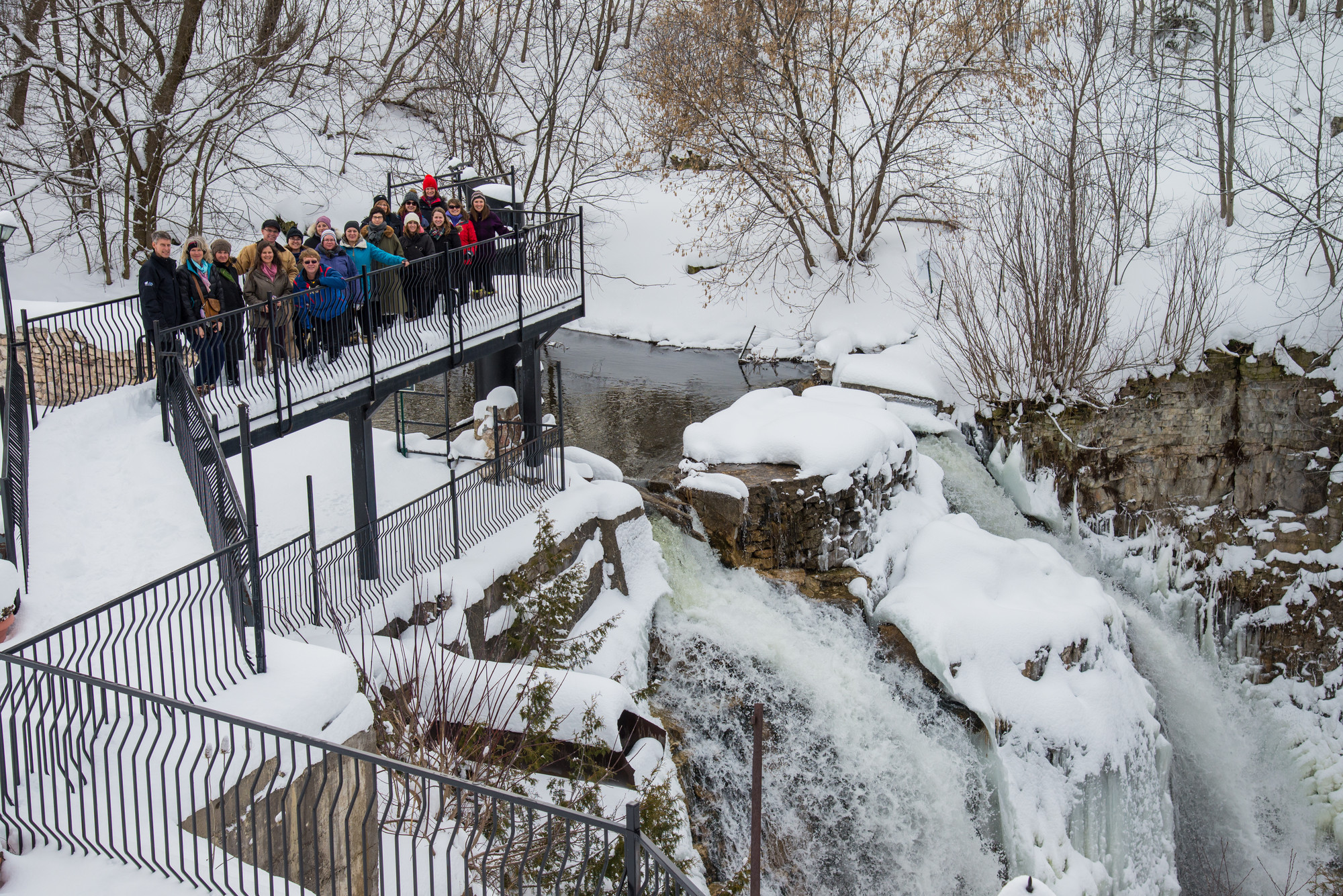 Group of people standing on a metal viewing platform above a partially frozen waterfall, surrounded by snowy trees and rock cliffs.