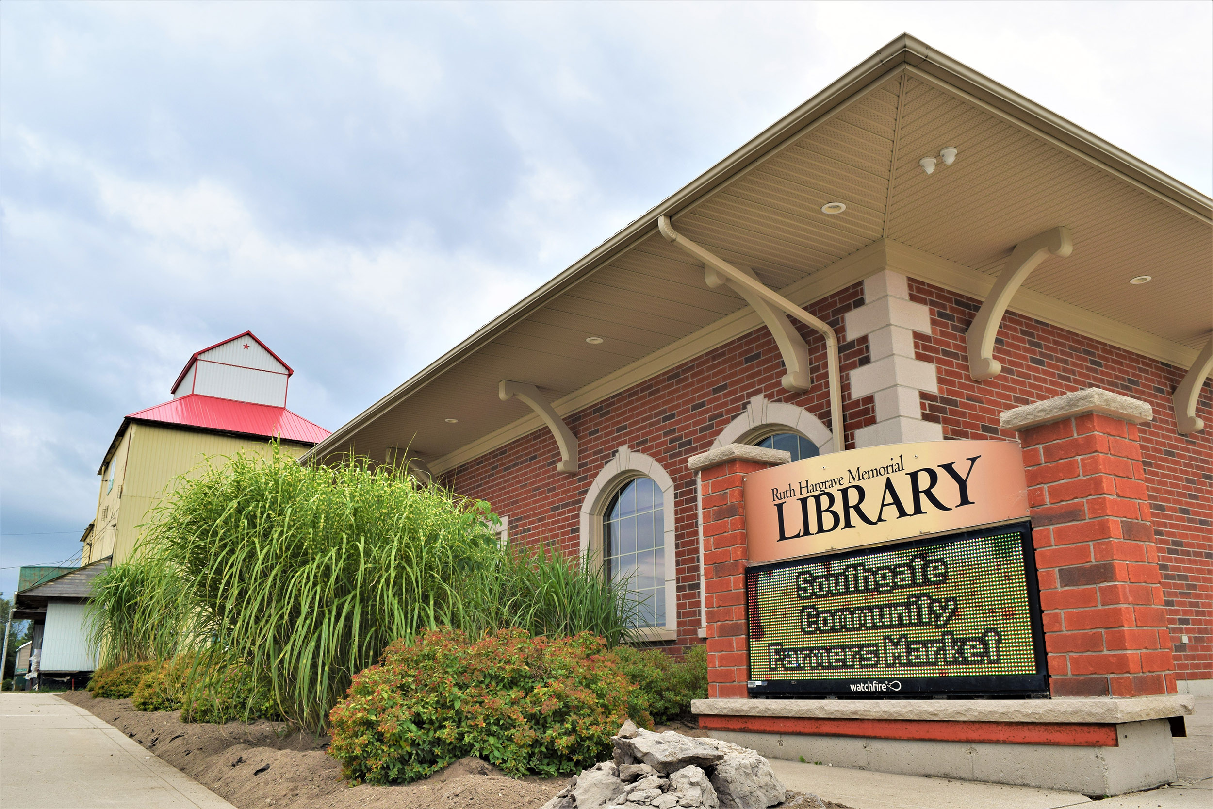 Brick library building with arched windows and a sign out front beside tall grasses.