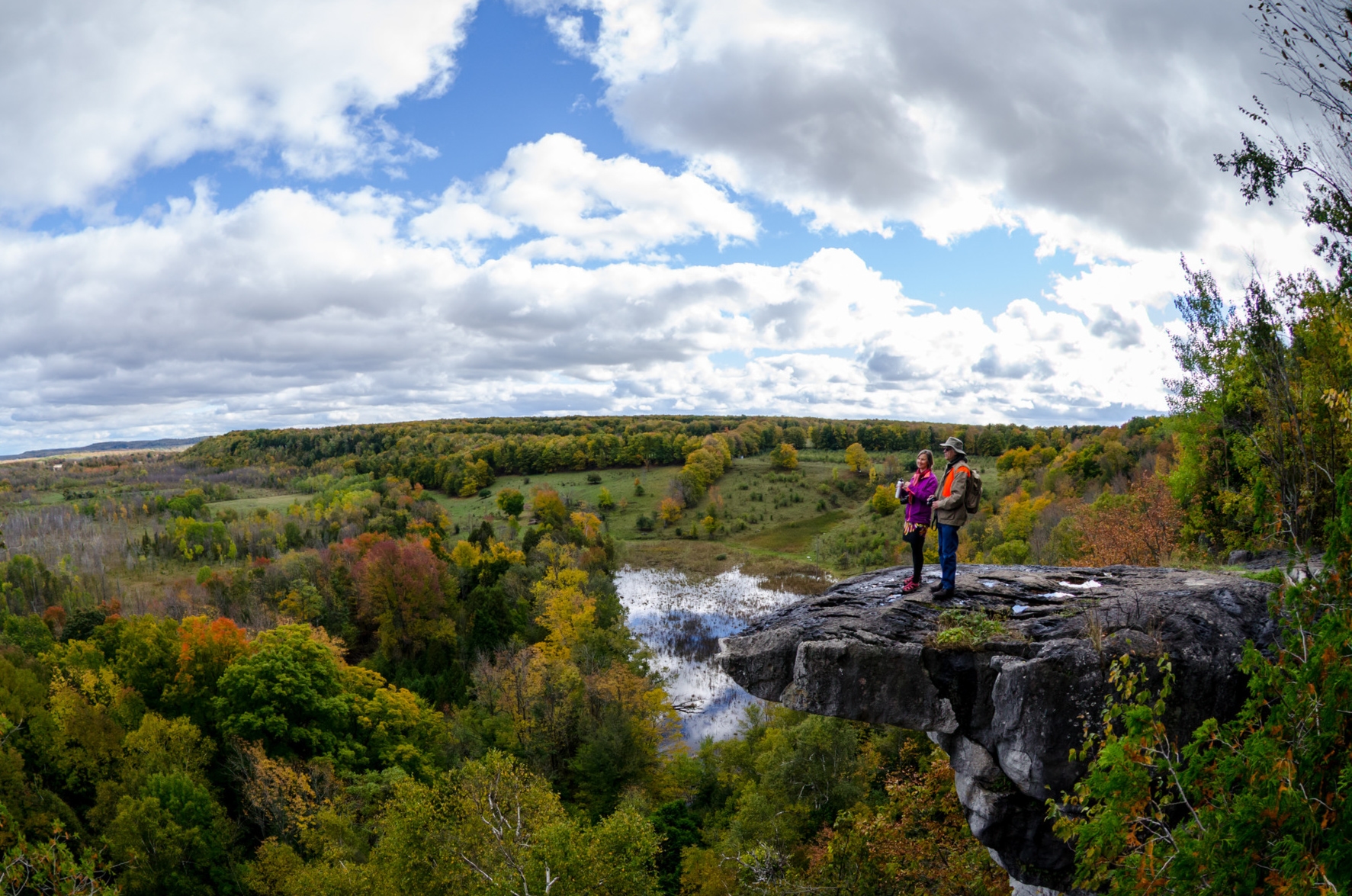 On a cloudy day, two hikers stand atop a rocky outcrop high above a scene below featuring trees, a pond, and fields.