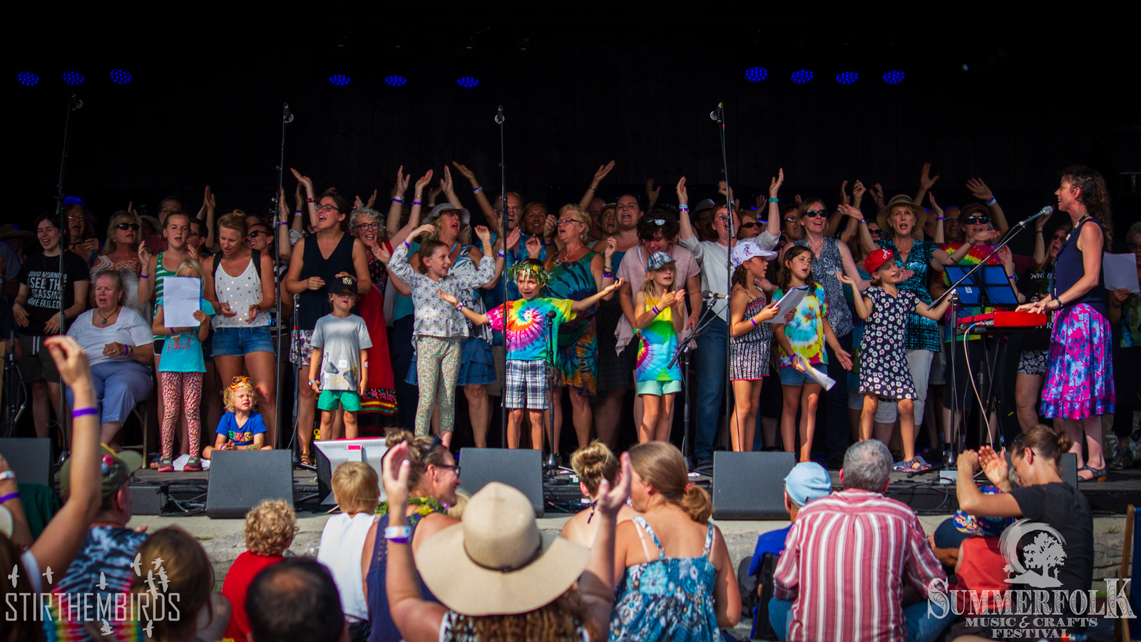 A large group of people singing, with hands raise, on a stage before an audience.