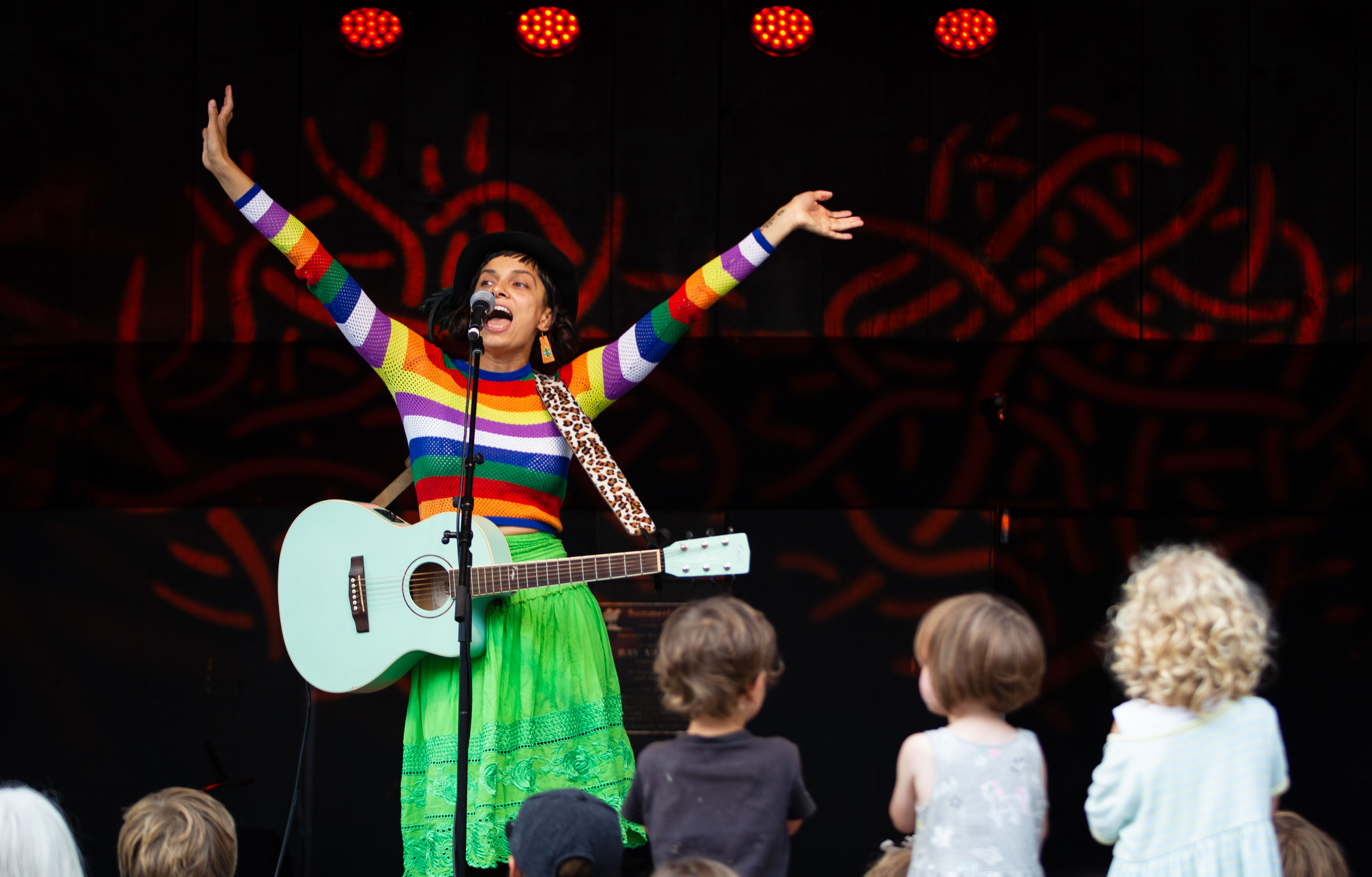 A performer with an acoustic guitar lifts her hands up as children watch closely.