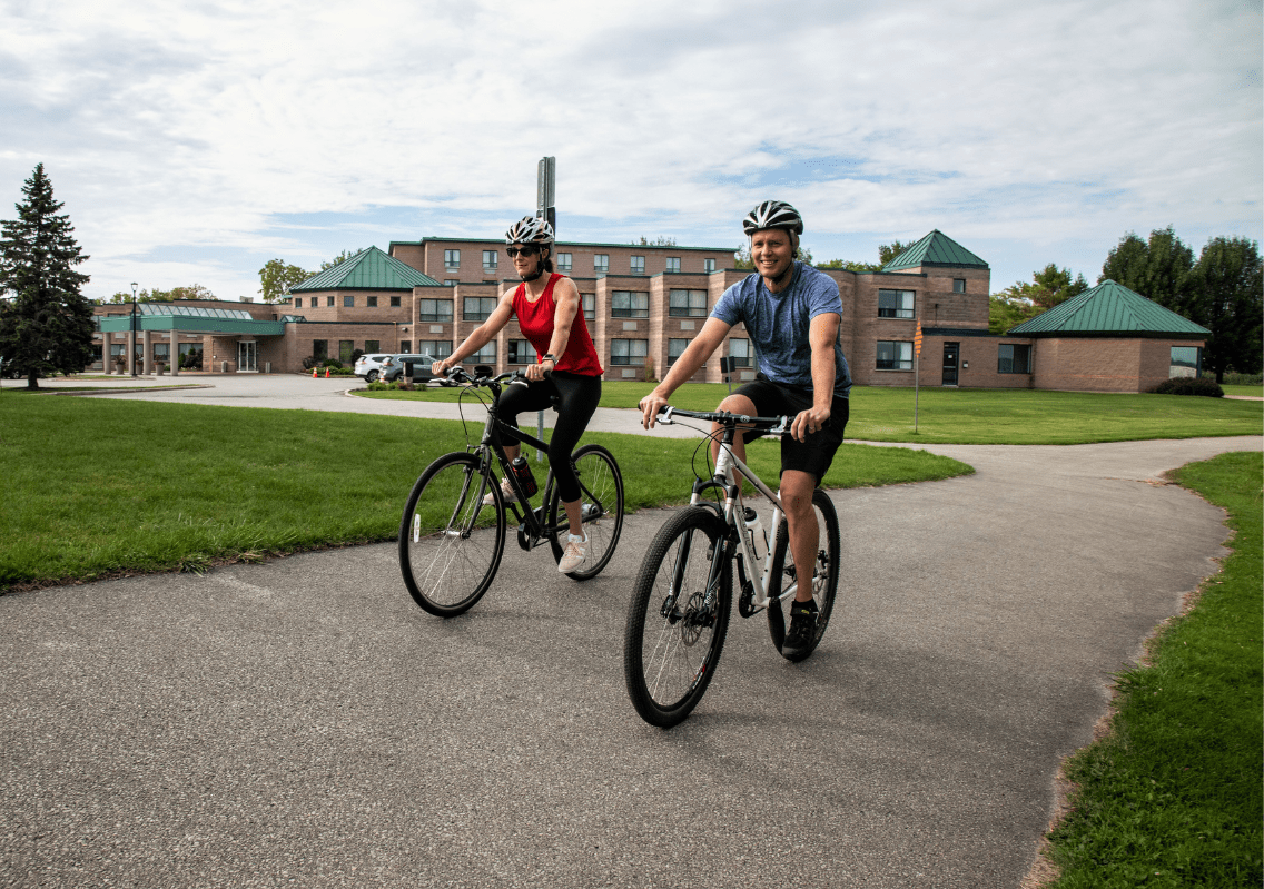 Two cyclists ride along a paved trail in front of a hotel building.