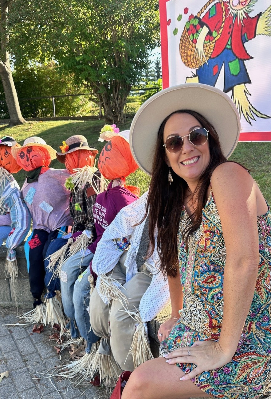 A smiling woman in a hat sits beside a line of scarecrows.