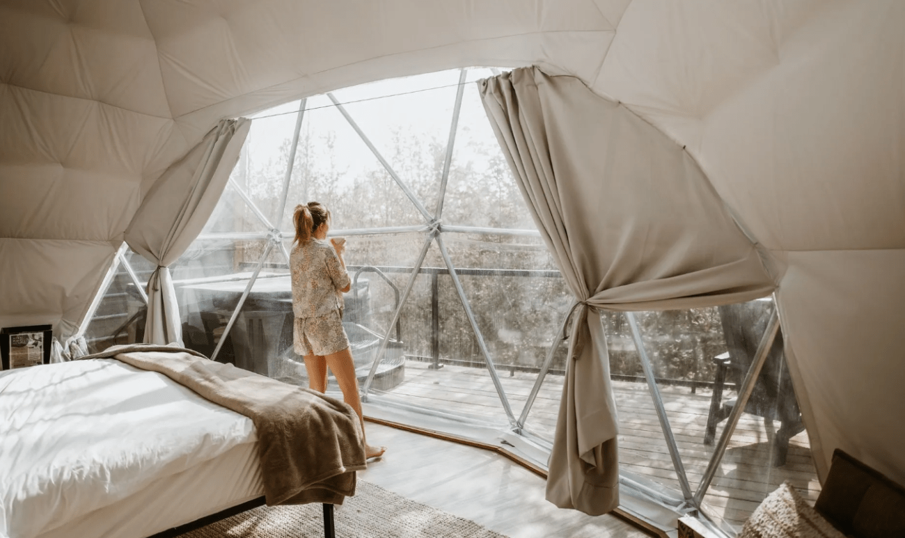 interior of a geodesic camping dome, a woman looks out at the view while drinking coffee