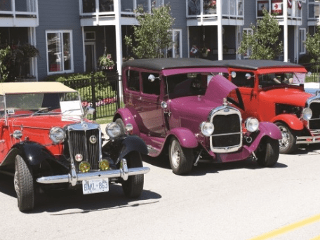 Three antique cars in front of a row of blue houses.