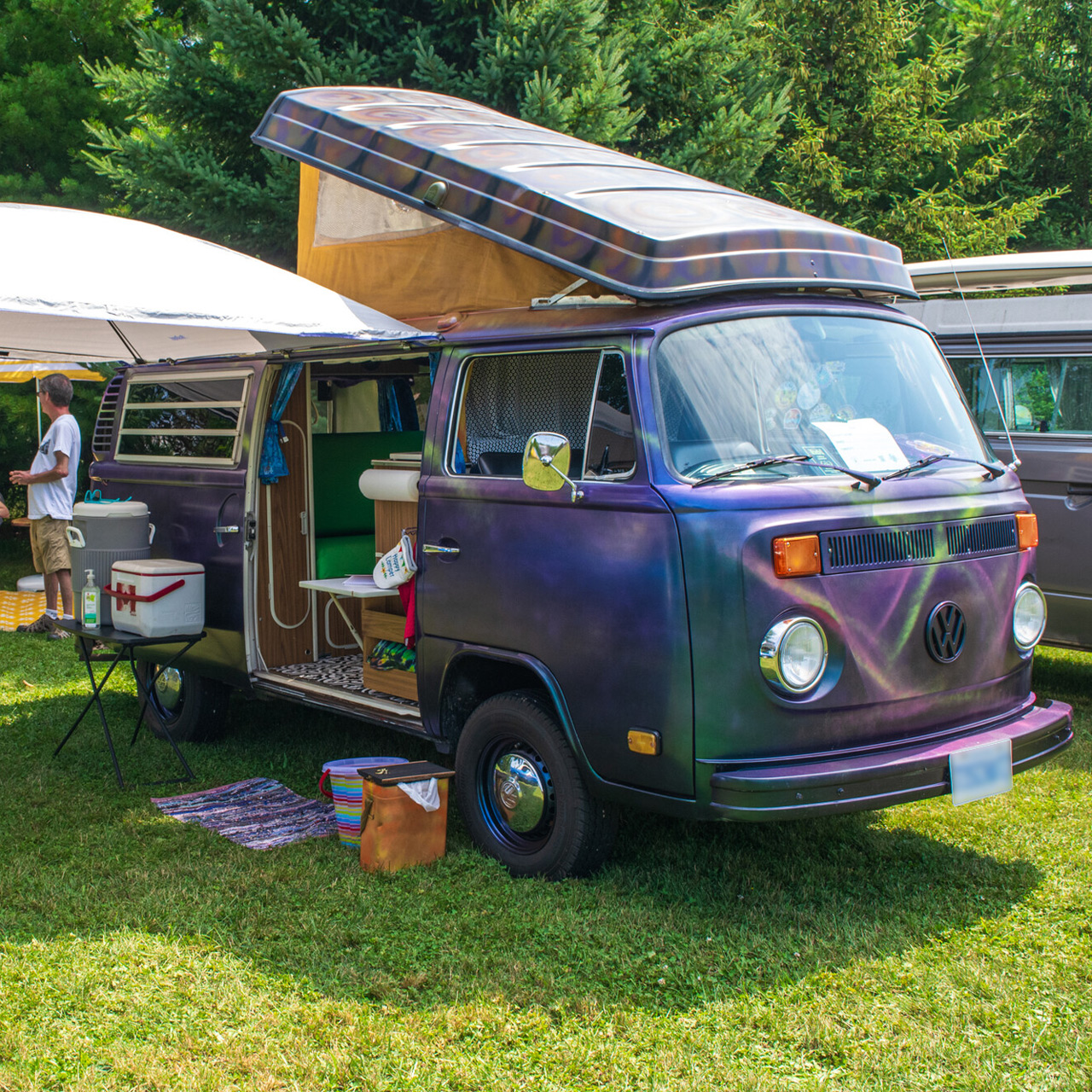 Vintage camper van set up at a grassy campsite, with an open side door, awning, and camping gear among trees.