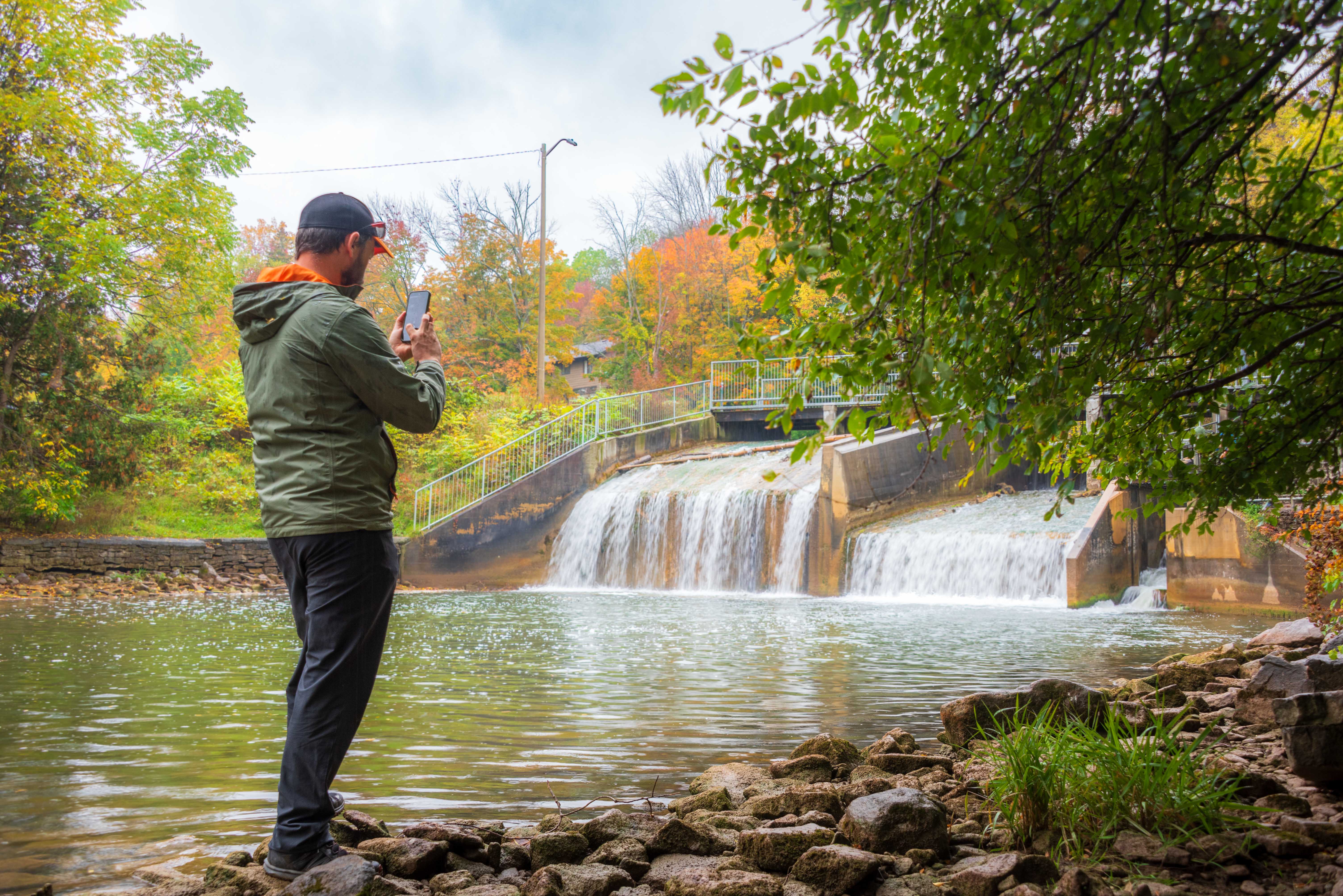 A person stands beside a river, taking a photo of cascading water at a dam.