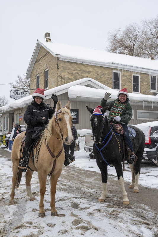 Two people on horses waving at the camera on a snowy street