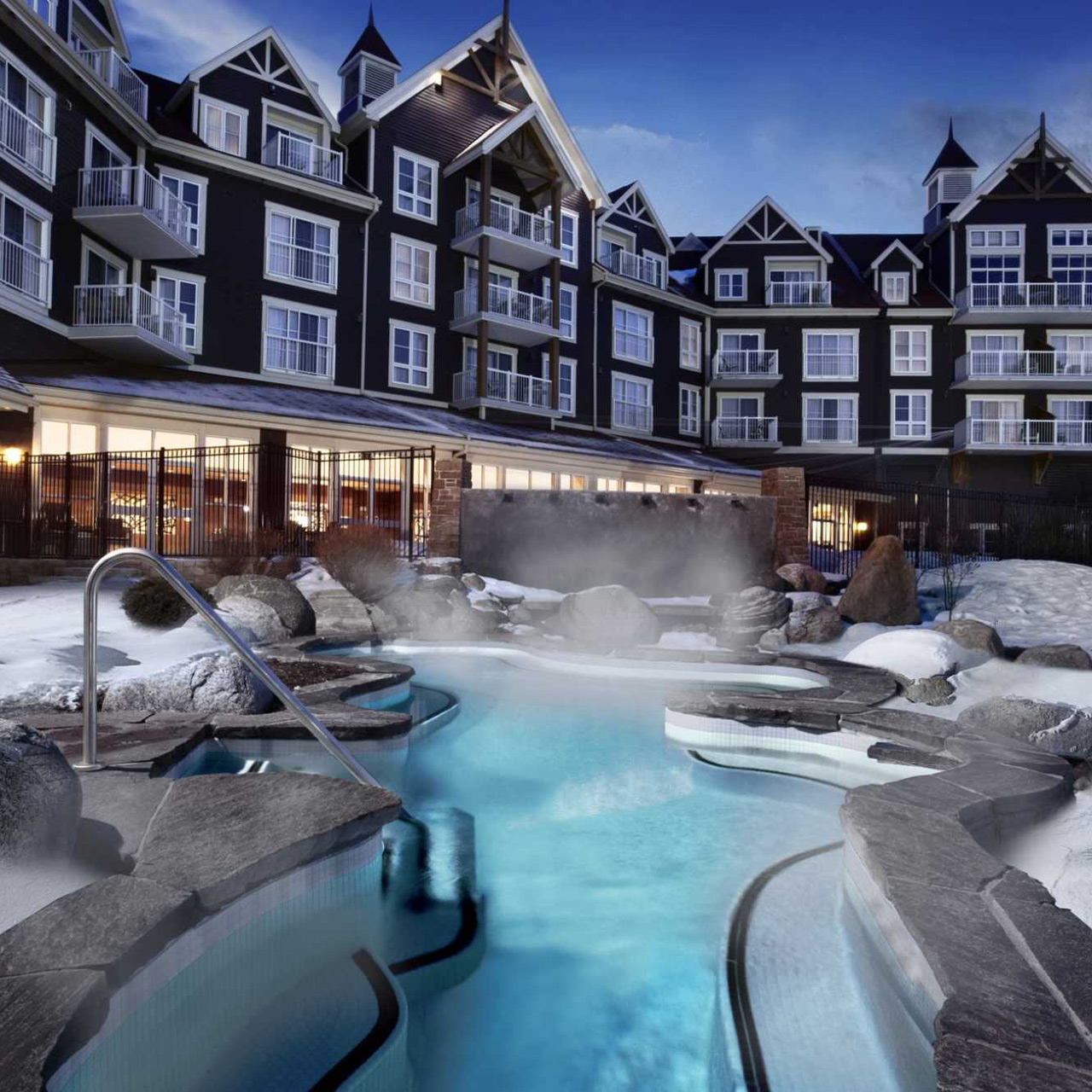Steaming outdoor pool at a resort hotel at dusk, surrounded by stone, snow, and chalet‑style buildings.