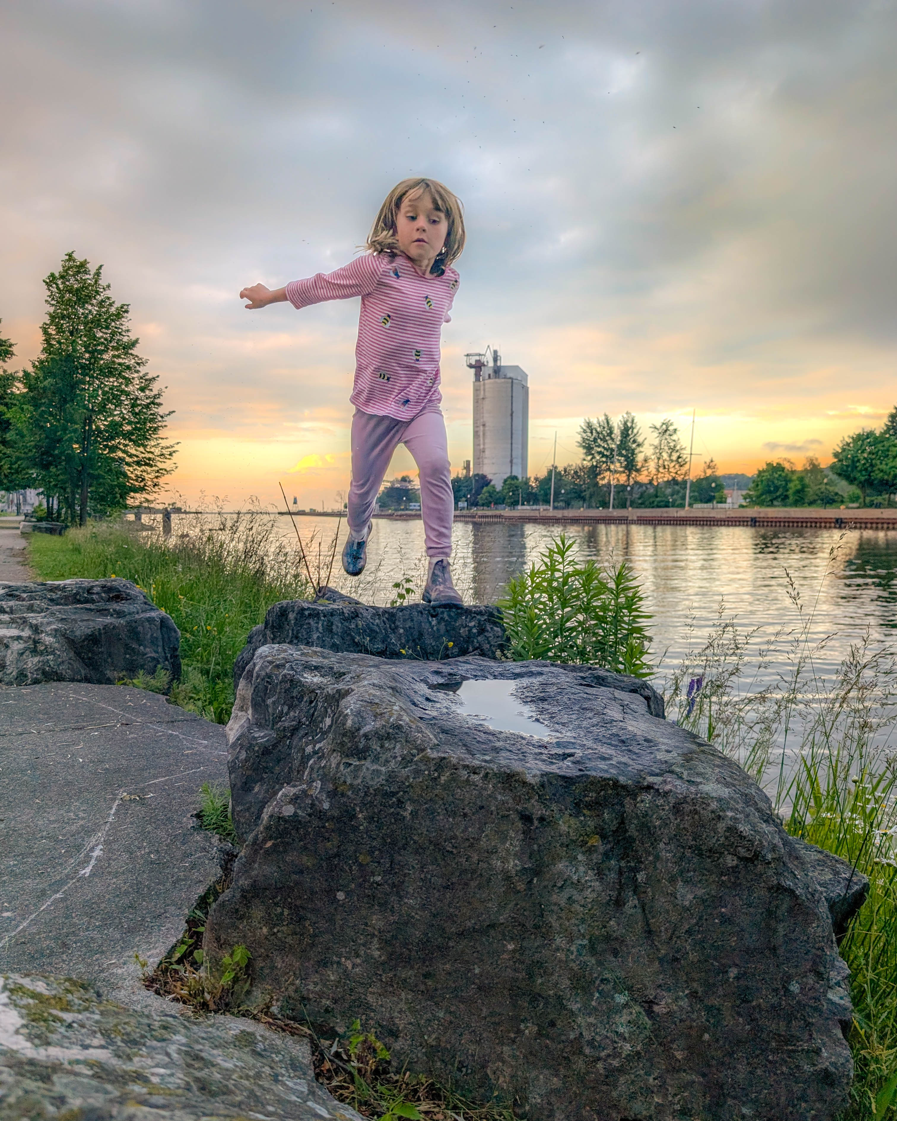 A child jumps between large stones beside a harbour.