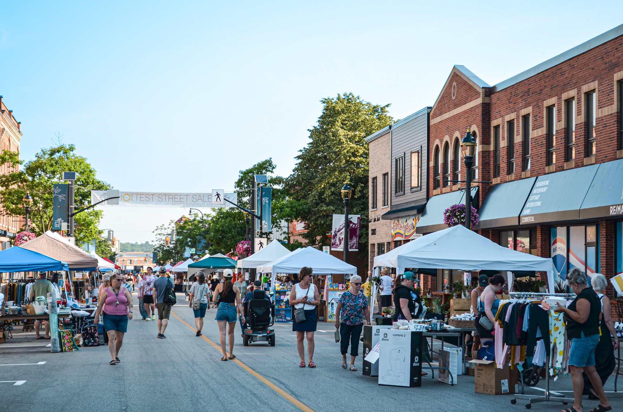 Busy downtown street market with vendor tents lining the road, shoppers browsing stalls, and brick storefronts under a clear sky.