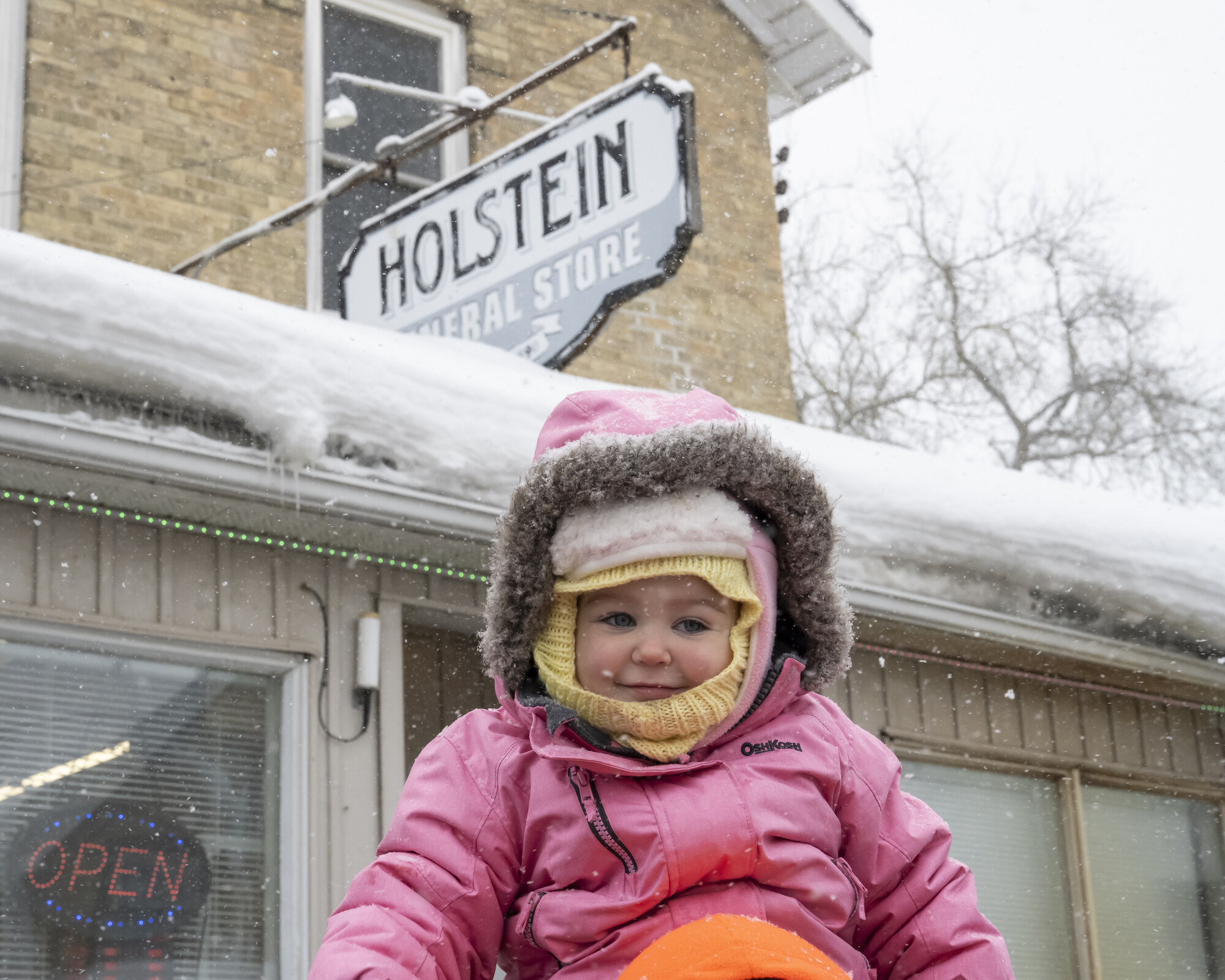 An infant wears a pink snowsuit on a snowy day in front of the Holstein General Store.