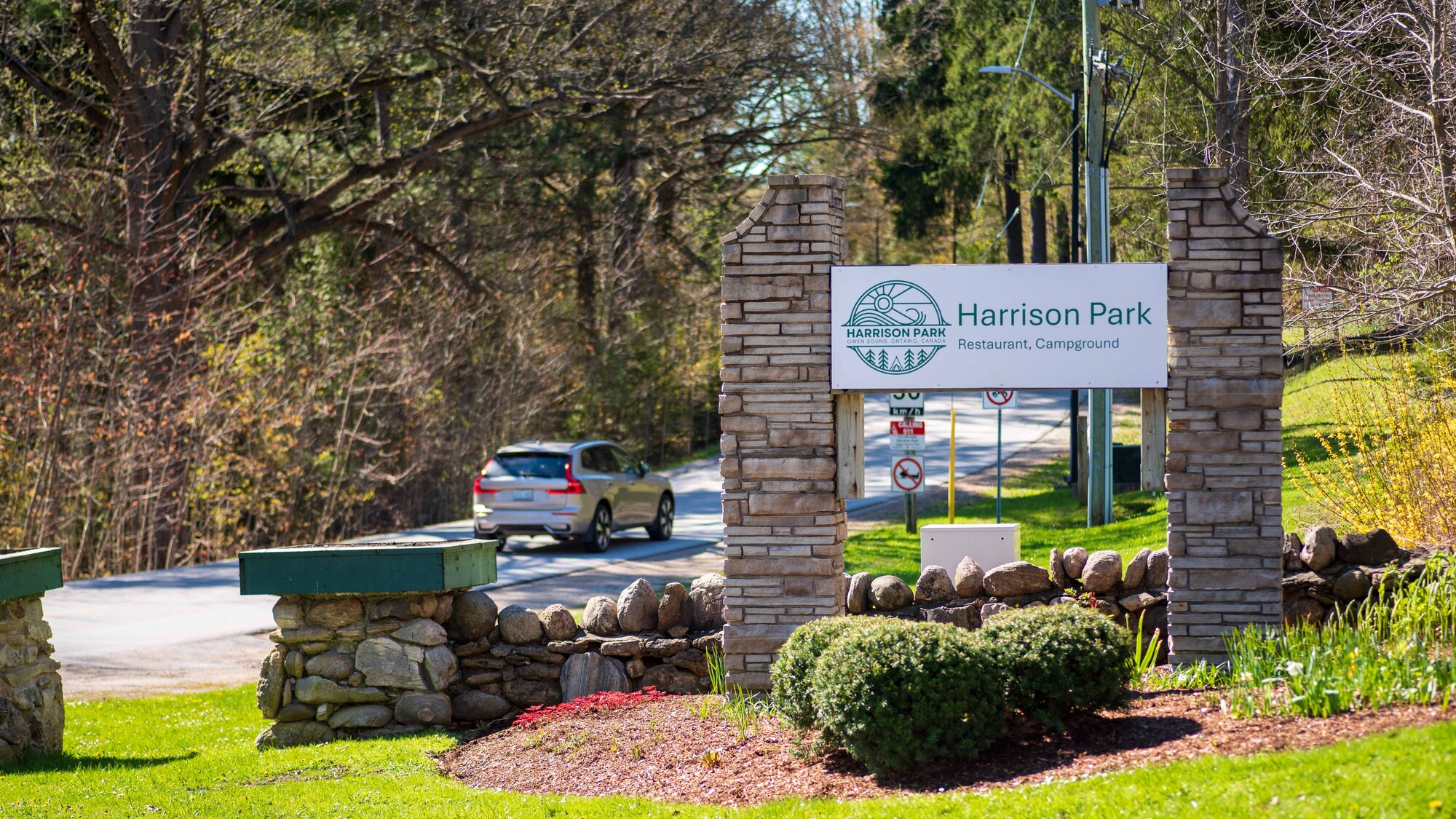 The entrance to Harrison Park. A vehicle drives past a sign and flowerbeds.