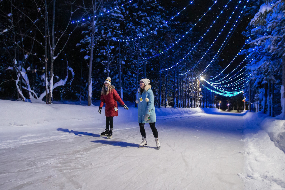 Two people skating along a path in the forest with blue string lights above them.
