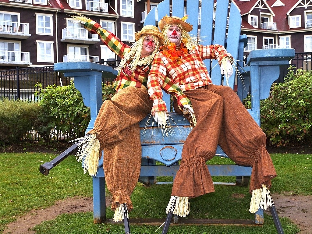 Two people dressed as scarecrows, wearing stilts, sit on an oversized blue chair.