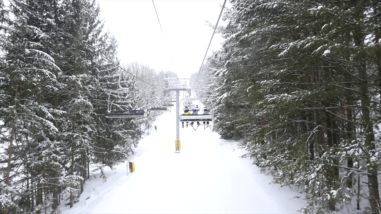 Rider view from an operating chairlift moving through snowy trees.