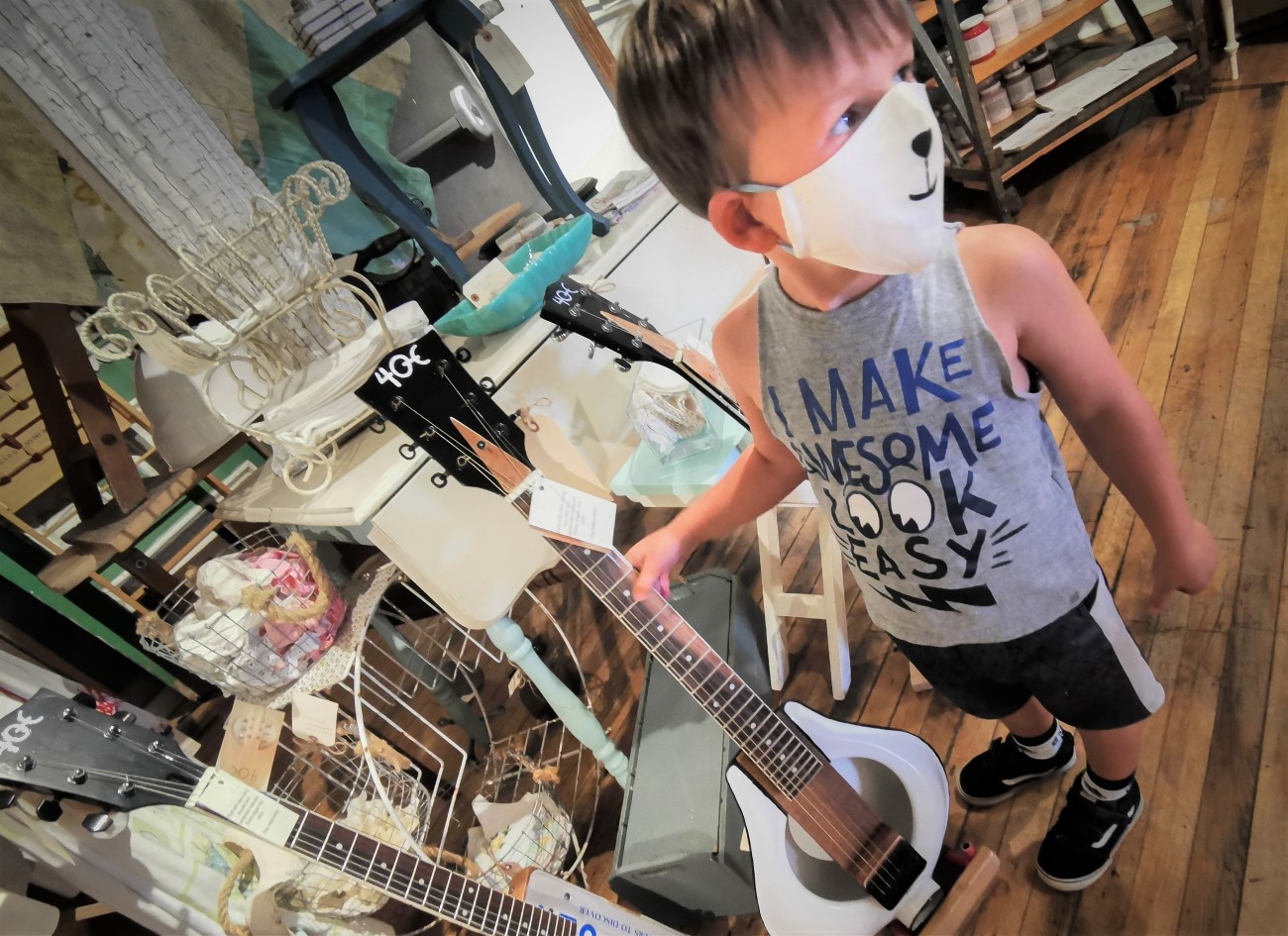 A child wearing a facemask runs his thumb across the strings of a guitar.