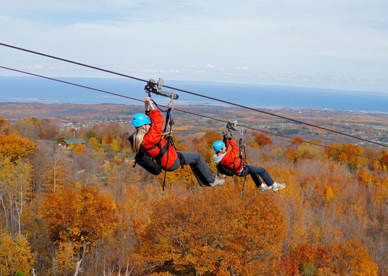 Two people on a zipline above trees displaying fall colours.