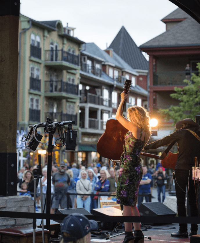 On an outdoor stage, a performer lifts up an acoustic guitar before a crowd.
