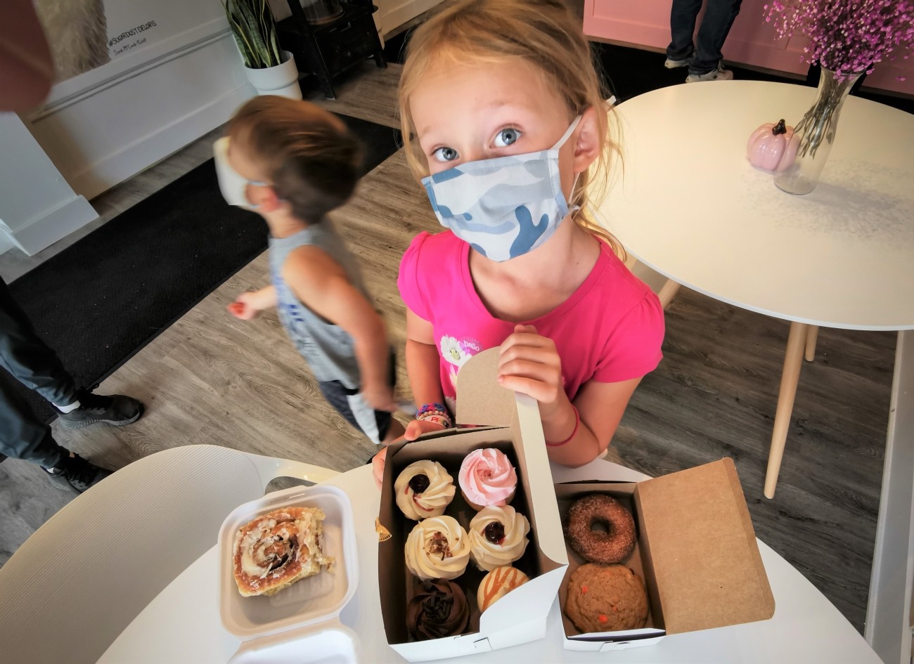 A child wearing a facemask holds open a box of cupcakes.